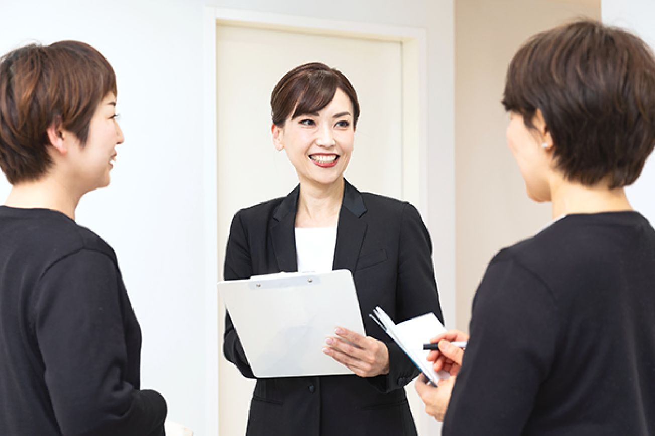 手にバインダーを持った水戸さんがスタッフ二人と向かい合って笑顔でミーティングしている画像。スタッフは手帳を開いている。